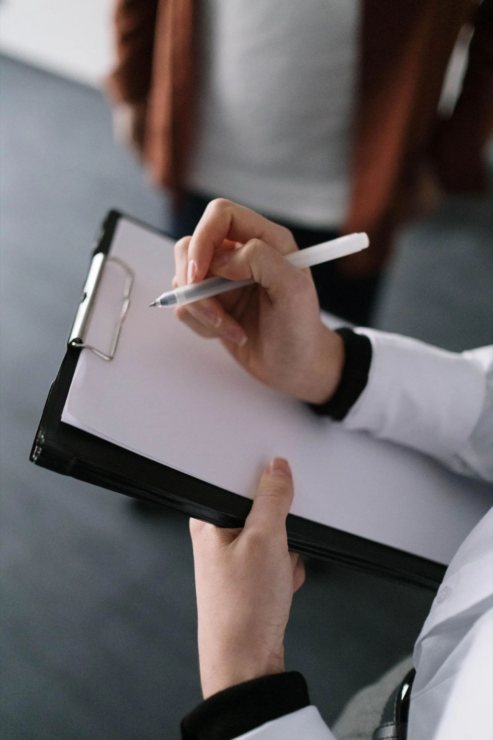 Anamnese: Eine Hand notiert mit dem Stift auf einem Klemmbrett, im Hintergrund eine Person